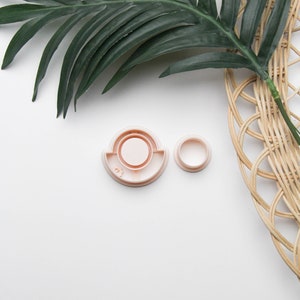 May include: Two light pink, circular cookie cutters. One is a larger, rounded rectangle shape with a circle inside. The other is a smaller circle. A green palm leaf and woven wicker basket are in the background.
