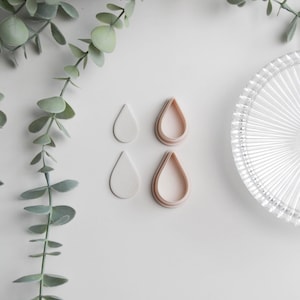 May include: Overhead shot of four teardrop-shaped earring components. Two are solid white, and two are light pink with a teardrop outline. A clear, ridged glass plate and green leafy plant are also in the image.