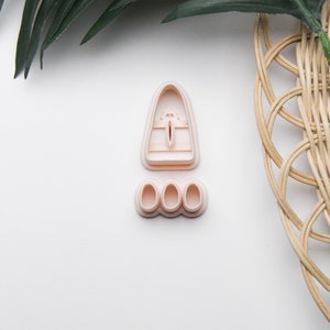 May include: Two light pink cookie cutters. One is a tall, rounded shape with a vertical slit and horizontal lines. The other is a set of three small, oval shapes. The items are on a white surface with green leaves and a woven basket.