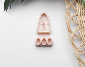 May include: Two light pink cookie cutters. One is a tall, rounded shape with a vertical slit and horizontal lines. The other is a set of three small, oval shapes. The items are on a white surface with green leaves and a woven basket.