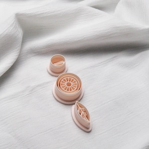 Peut inclure: Ensemble de découpoirs à biscuits rose clair de différentes formes, dont un cercle avec un motif ensoleillé, un petit cylindre et une feuille. Les découpoirs sont disposés sur un fond de tissu blanc texturé.