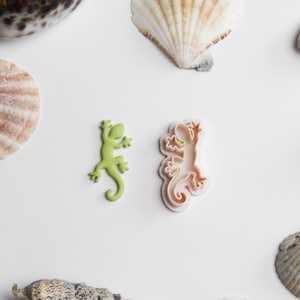 May include: Two plastic cookie cutters shaped like lizards, one green and one pink. The cutters are on a white surface with seashells.