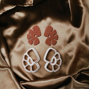 May include: Two sets of craft cutters. The top set is a reddish-brown leaf shape with a textured surface. The bottom set is a white, open-design seashell shape. The cutters are on a brown satin fabric.