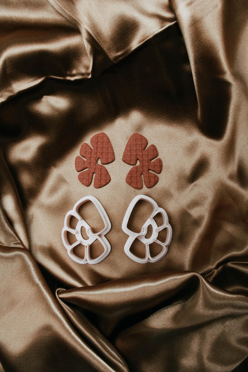 May include: Two sets of craft cutters. The top set is a reddish-brown leaf shape with a textured surface. The bottom set is a white, open-design seashell shape. The cutters are on a brown satin fabric.