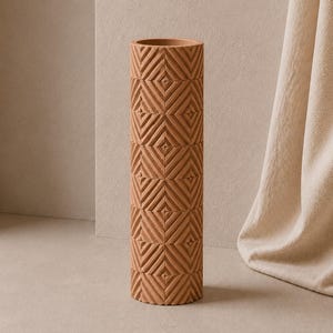 May include: A tall, cylindrical terracotta-coloured vase featuring a repeating geometric pattern of embossed diagonal lines and small squares. The surface has a matte finish with a consistent earthy tone throughout the entire structure.