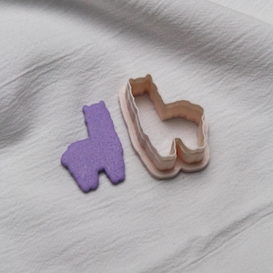 May include: A purple llama-shaped cookie cutter and a purple llama-shaped cookie on a white surface. The cookie cutter is light pink with a beige outline. The cookie is a solid purple color.