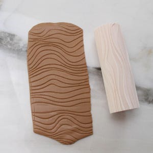 May include: A brown clay slab with a wood grain pattern and a light-colored wooden rolling pin. The slab is rectangular with rounded edges and the rolling pin is cylindrical. The wood grain pattern is visible on both items.