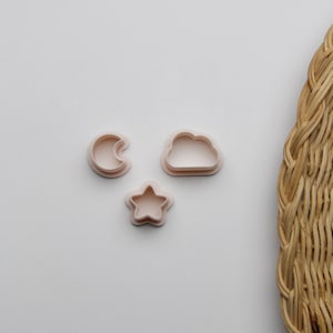 May include: Three plastic cookie cutters in the shape of a crescent moon, a cloud, and a star. The cutters are on a white surface.