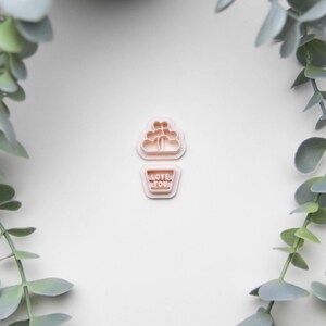 May include: Two light pink cookie cutters. The top cutter is shaped like a cloud with hearts. The bottom cutter is a rectangle with the words "LOVE YOU" in capital letters. Green leafy plants frame the image.