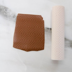 May include: A brown, textured clay piece and a white roller with a similar pattern are displayed on a white surface. The clay piece has a rectangular shape with a woven-like texture. The roller is cylindrical and also has a woven pattern.