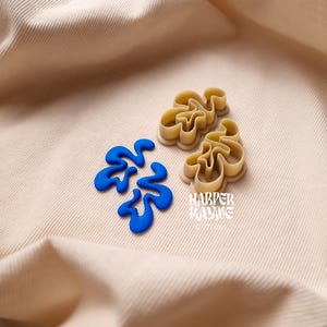 May include: Two beige plastic cookie cutters shaped like abstract swirls, one with blue frosting inside. The cutters are on a beige fabric background with the text "Harper Kayne" in a retro font.