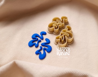 May include: Two beige plastic cookie cutters shaped like abstract swirls, one with blue frosting inside. The cutters are on a beige fabric background with the text "Harper Kayne" in a retro font.