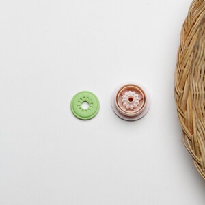 May include: Two round plastic buttons, one green and one pink. The green button has a flower design in the center. The pink button has a flower design with a smaller circle in the center.