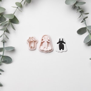 May include: Three ghost-shaped cookie cutters in pink and black, arranged on a white surface. The cutters are in different sizes and designs, with one black and white ghost. Green foliage frames the top of the image.