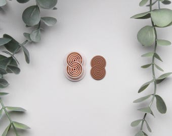 May include: Two polymer clay cutters in the shape of the number eight, one light pink and one brown. The cutters have a spiral design inside. The background is white with green eucalyptus leaves.