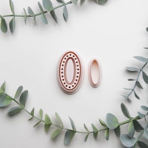 May include: Two pink plastic cookie cutters in the shape of an oval. The larger cutter has a decorative border with small circles. The smaller cutter is plain.