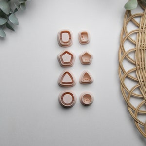 May include: A set of eight pink plastic cookie cutters in various shapes, including a square, circle, pentagon, triangle, and heart. The cutters are arranged in two rows of four on a white surface.