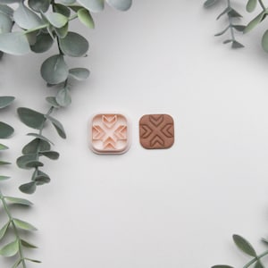 Op de afbeelding: Een close-up van twee vierkante koekjesvormen. De vorm aan de linkerkant is roze en heeft een geometrisch ontwerp. De vorm aan de rechterkant is bruin en heeft hetzelfde ontwerp, maar is gegraveerd.