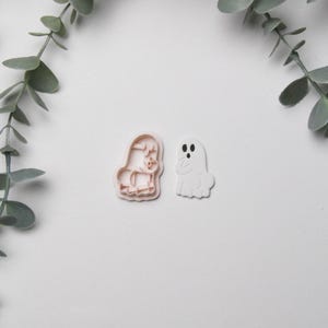May include: Two Halloween-themed cookie cutters on a white surface. One is a light pink outline of a ghost, and the other is a light pink outline of a witch. Green leafy branches are in the background.
