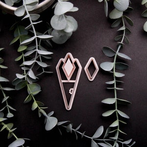 May include: A set of two pink plastic cookie cutters in the shape of a diamond and a stylised torch. The cutters are on a black background with green eucalyptus leaves.