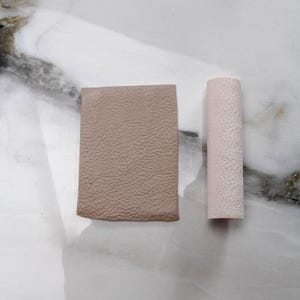 May include: Two small, textured objects. One is a rectangular piece of tan material. The other is a cylindrical object in a light pink color. Both objects have a similar textured surface.
