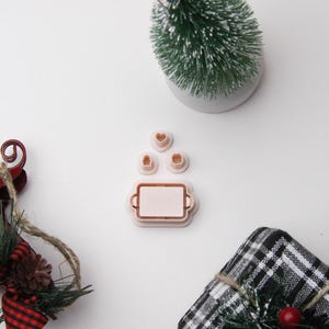 May include: A festive arrangement featuring a miniature Christmas tree with a star topper, alongside a set of pink cookie cutters in the shapes of a tray and hearts. A wrapped gift and decorative accents complete the holiday theme.