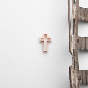 May include: A light pink cross-shaped cookie cutter. The cutter has a simple, classic design with a cross shape inside a slightly larger outline. The background is white, with a rustic wooden ladder on the right side.