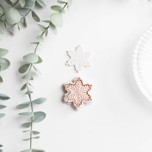 May include: Two snowflake-shaped decorations, one white and one light brown with a detailed design, rest on a white surface. Green leafy branches are in the upper left corner. A decorative plate is in the upper right corner.