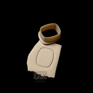 Peut inclure: Un emporte-pièce en plastique beige en forme de rectangle arrondi. L'emporte-pièce est placé sur un morceau de fondant beige avec la même forme découpée. Le fondant porte l'inscription "HARPER BAYNE CUTTERS".