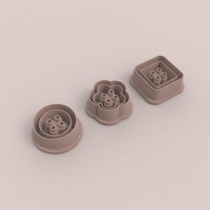 May include: Three dusty rose coloured cookie cutters in circular, flower, and square shapes. Each cutter features a raised outer rim and a central four-hole plunger mechanism designed for creating embossed patterns on dough or clay surfaces.