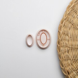 May include: Two pink plastic cookie cutters, one shaped like a circle with an oval inside, and the other shaped like an oval. The cutters are on a white surface.