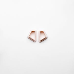 Op de afbeelding: Twee lichtroze geometrisch gevormde oorbellen met een holle binnenkant, tegen een witte achtergrond. De oorbellen hebben een plat, hoekig ontwerp met een licht afgeronde rand. De algehele esthetiek is minimalistisch en modern.