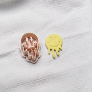 May include: A light pink cookie cutter and a yellow cookie. The cutter is shaped like a melting smiley face with lightning bolt eyes. The yellow cookie is a melting smiley face.