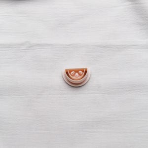 Peut inclure: Emporte-pièce rose clair en forme de demi-cercle avec un motif de pastèque. Le découpoir a un bord arrondi et trois petites indentations circulaires à l'intérieur de la forme de la pastèque. Sur une surface blanche texturée.