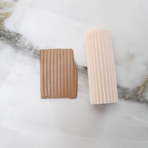 May include: Two textured clay pieces. One is a rectangular shape in a brown hue, with vertical ridges. The other is a cylindrical shape in a light pink color, also with vertical ridges. Both pieces are on a white marble surface.