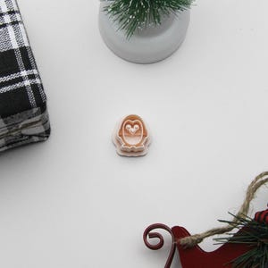 Könnte beinhalten: Ein hellbrauner Pinguin-Ausstecher. Das Bild zeigt auch einen kleinen grünen Weihnachtsbaum, einen roten Schlitten-Ornament und ein schwarz-weiß kariertes Tuch, alles vor weißem Hintergrund.