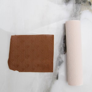 May include: A brown rectangular clay piece and a light pink cylindrical roller, both with a repeating geometric pattern. The clay piece is textured and has a rough edge. The roller is smooth and cylindrical, with the same pattern.