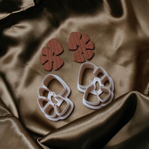 May include: Two pairs of leaf-shaped earrings and two leaf-shaped cookie cutters. The earrings are a textured, reddish-brown color. The cookie cutters are white. All items are on a brown satin surface.