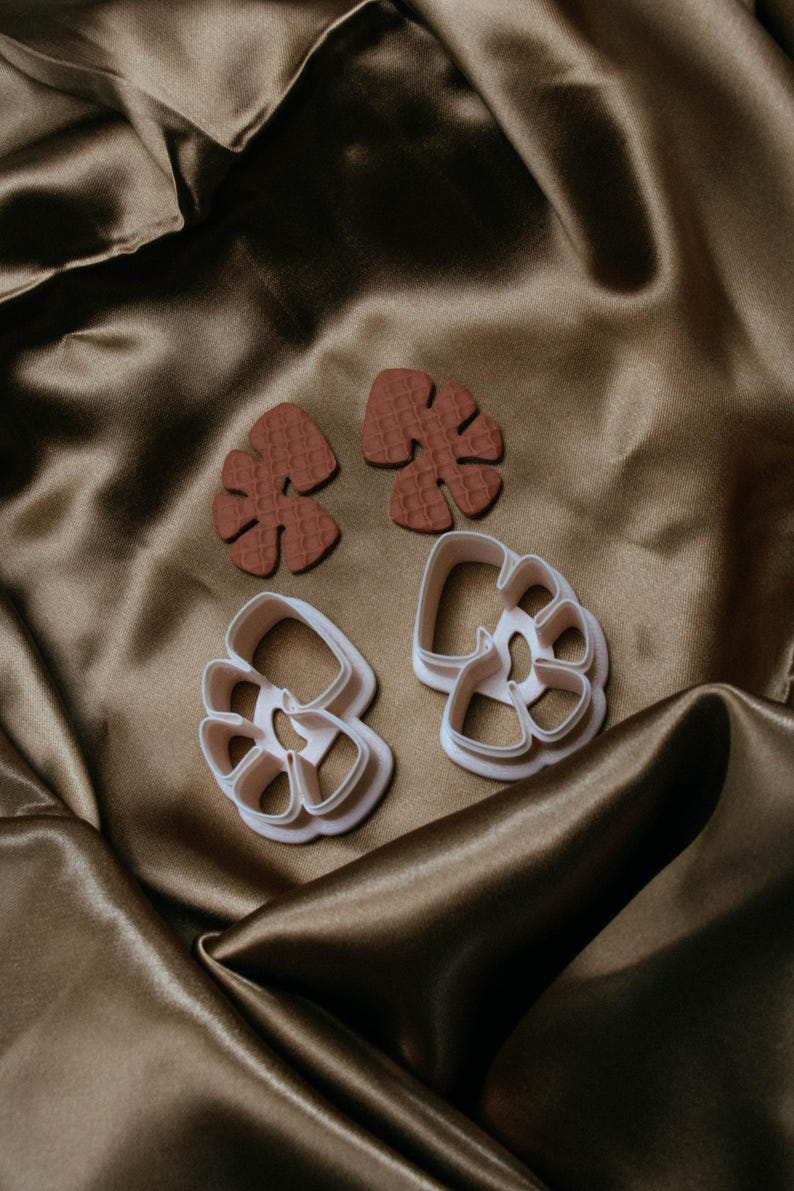 May include: Two pairs of leaf-shaped earrings and two leaf-shaped cookie cutters. The earrings are a textured, reddish-brown color. The cookie cutters are white. All items are on a brown satin surface.