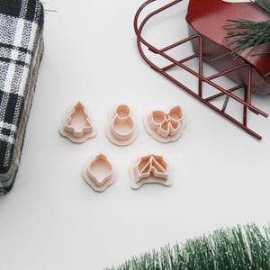 May include: Five light pink cookie cutters in festive shapes: a Christmas tree, snowman, bow, ornament, and holly. A red metal sleigh and a small artificial Christmas tree are also visible.