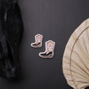 Peut inclure: Deux emportes-pièces en forme de bottes de cowboy en plastique rose sur un fond noir.