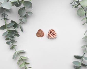 May include: Two small, brown, circular stamps with a Celtic knot design are displayed on a white surface. The stamps are surrounded by green eucalyptus leaves, creating a natural frame. The stamps are different sizes.