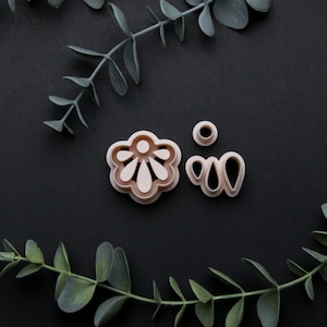 May include: A set of four cookie cutters in a light pink color. The cutters are shaped like a flower, two leaves, and a circle. The cutters are on a black background.