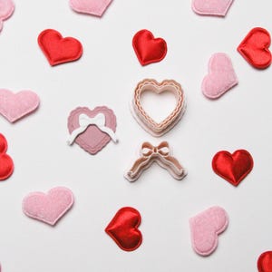Può includere: Una vista dall'alto mostra decorazioni a forma di cuore rosse e rosa e tagliapasta sparsi su uno sfondo bianco. I cuori sono fatti di feltro e raso. Sono visibili due tagliapasta, uno a forma di cuore e uno a forma di fiocco.