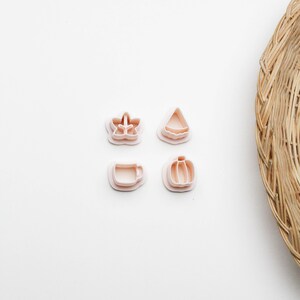 Peut inclure: Quatre emporte-pièces roses pâles de différentes formes : une fleur, un triangle, une tasse et une citrouille. Ils sont disposés sur une surface blanche, avec un panier en osier en arrière-plan. Idéal pour la pâtisserie et le bricolage.