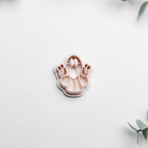 May include: A light pink ghost-shaped cookie cutter with a friendly face and outstretched arms. The cutter is made of plastic and is designed for making Halloween-themed cookies. The image is set against a white background with green foliage.