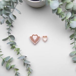 May include: Two heart-shaped cookie cutters. One is a larger heart with a decorative sunburst design, and the other is a smaller, plain heart. The cutters are light pink and sit on a white surface, with green leafy branches in the background.