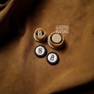 May include: Two white and black 8 ball cookie cutters and two finished 8 ball cookies on a brown suede surface. The cookie cutters are made by Harper Bayne Cutters.