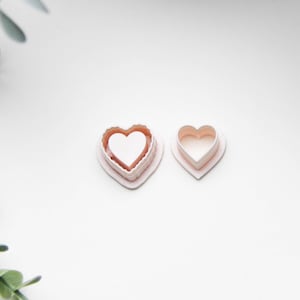 May include: Two pink heart-shaped cookie cutters on a white surface. One cutter has a scalloped edge, and the other is a simple heart shape. The cutters are likely used for baking or crafting.
