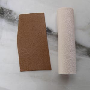 May include: A brown rectangular piece and a white cylindrical object with a textured surface. The objects are displayed on a white and gray marble surface.